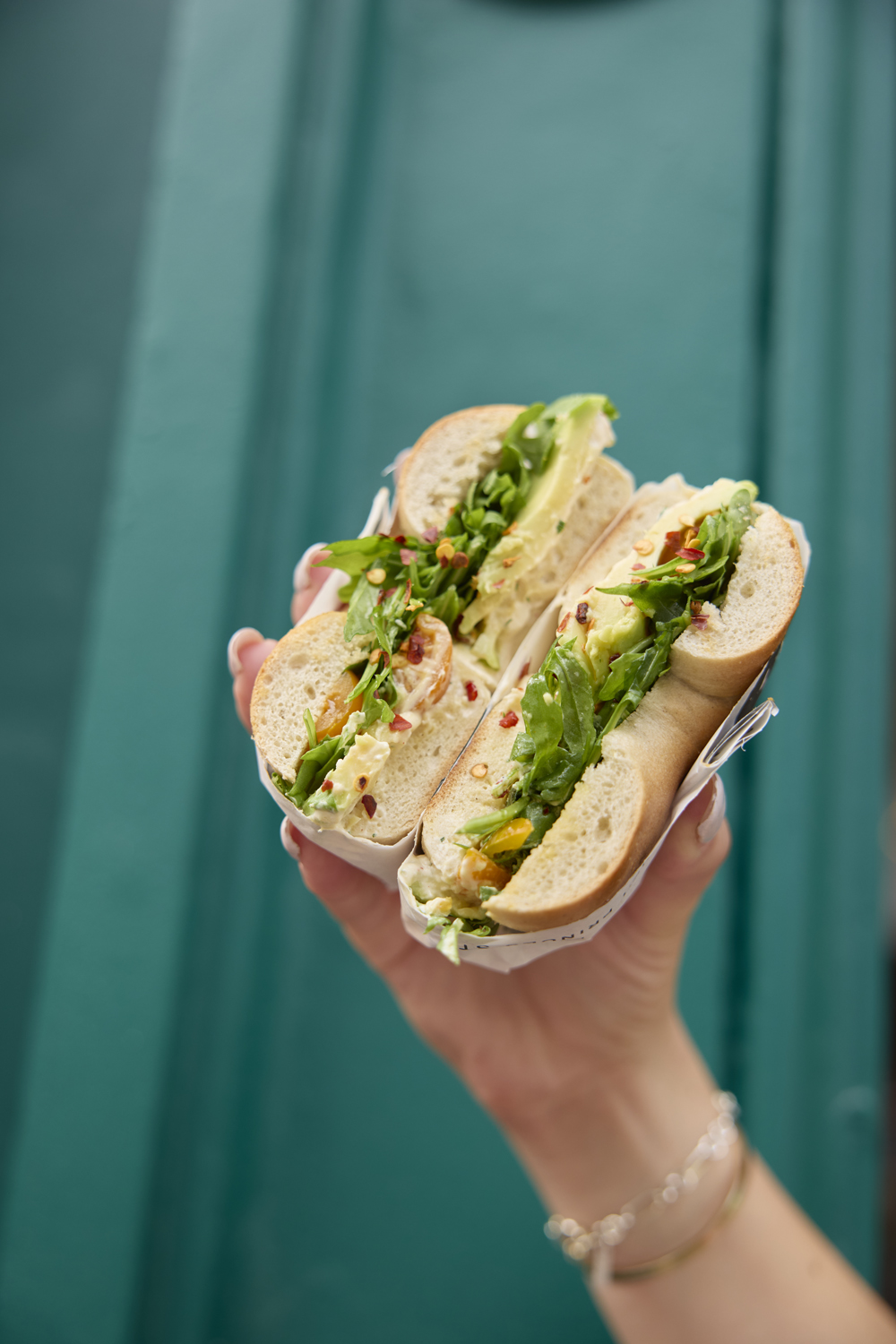 A wrapped bagel sandwich held up against a teal wall