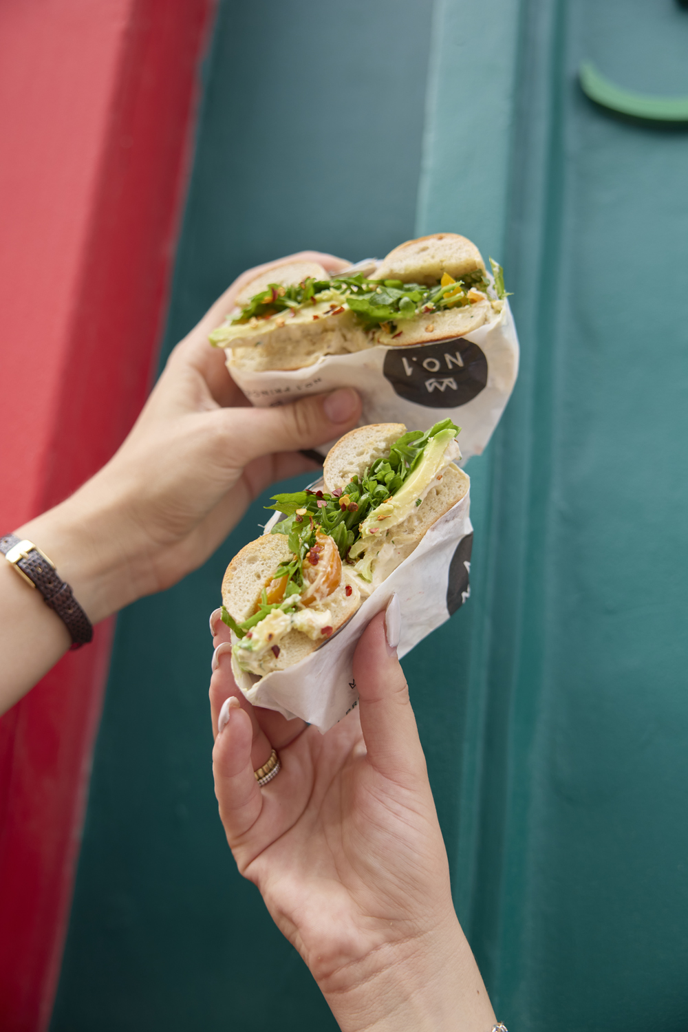 Hands holding two wrapped bagel sandwiches against a teal wall